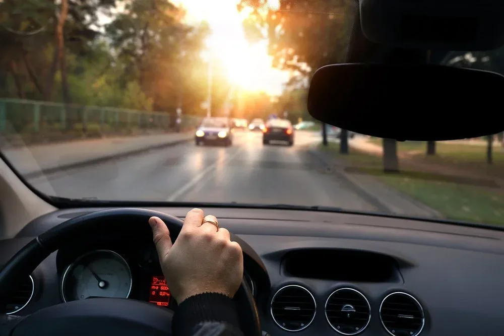 Driver's hand on steering wheel during sunset drive on tree-lined street with traffic ahead.