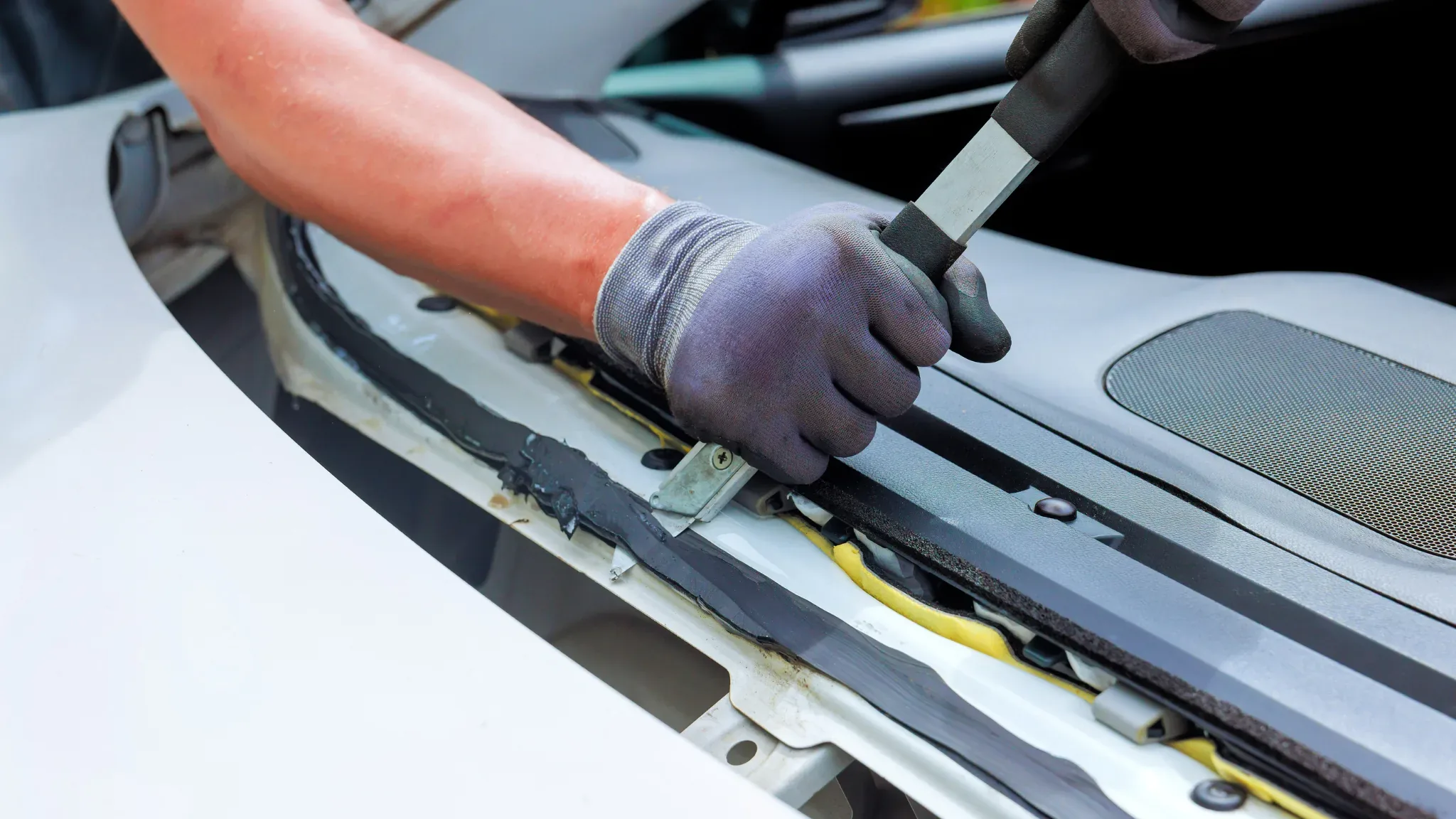 A gloved hand uses a flat metal pry tool to lift a black trim strip along the base of a white car's windshield, exposing yell
