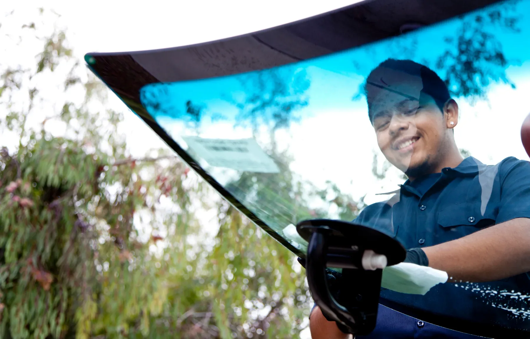 A smiling young man with short dark hair and an earring, wearing a dark navy polo shirt, holds a car windshield up by its suc