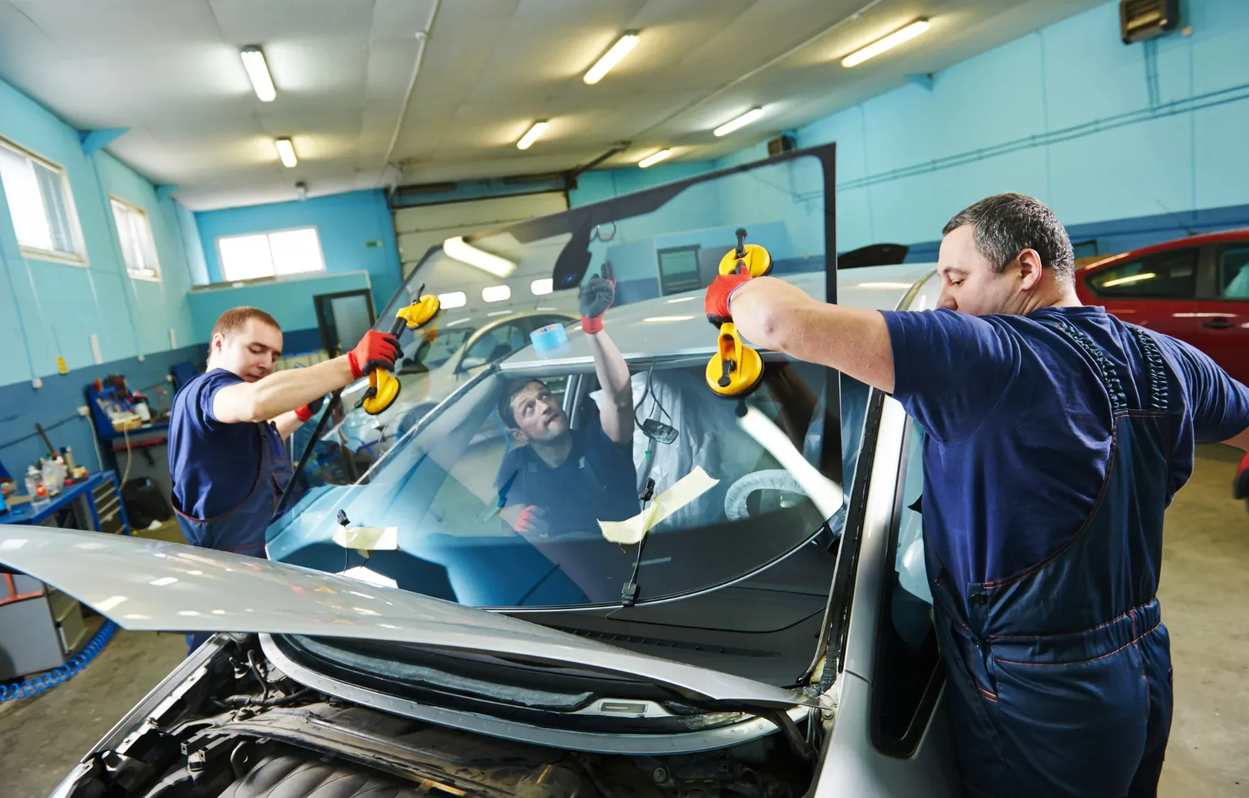 Two technicians in dark blue coveralls use yellow suction-cup handles to lift and position a new windshield onto a silver car